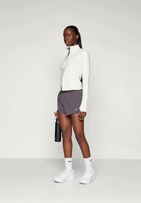 Hvid lynlåsjakke, grå shorts, hvide sportssko og en vandflaske. Glat tekstur, tætsiddende design og subtile brandaccenter.