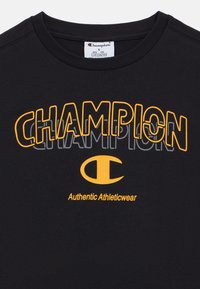 Μαύρο βαμβακερό μπλουζάκι με έντονο κίτρινο κείμενο "CHAMPION", κυκλικό λογότυπο και τη φράση "Authentic Athleticwear" στο κάτω μέρος.