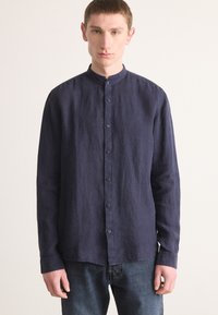 Marineblå langermet skjorte med mandarin krage, teksturert linestoff og knappestolpe; kombinert med denimjeans.