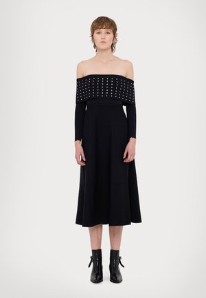 Zwarte off-shoulder jurk met een gestructureerde, gestickerde bodice en lange, aansluitende mouwen. Aansluitende rok met een golvende zoom, gecombineerd met zwarte enkelboots.