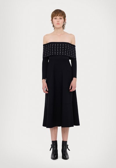 Vestido negro sin hombros con corsé texturizado y tachonado, y mangas largas y ajustadas. Falda acampanada con dobladillo en forma de ondas, combinada con botas negras hasta el tobillo.