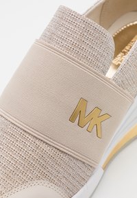 Gros plan sur une chaussure beige à enfiler avec une texture, dotée d'une bande élastique ornée du logo "MK" doré sur la partie supérieure.