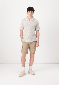 Camisa de botón de manga corta con un patrón zigzagueante en tonos pastel, combinada con pantalones cortos cargo beige que cuentan con bolsillos laterales. Zapatillas ligeras completan el look.