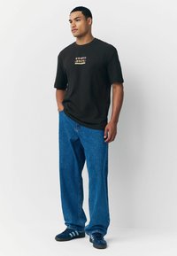 Schwarzes T-Shirt mit kurzen Ärmeln und buntem Grafikdetail auf der Brust, kombiniert mit lockeren blauen Jeans und blauen Sportschuhen.