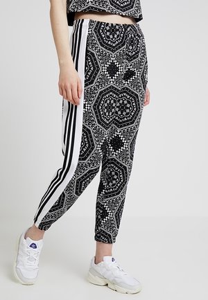 Femme portant un pantalon de jogging à motifs noir et blanc avec des bandes blanches sur les côtés et des baskets blanches avec un logo violet.