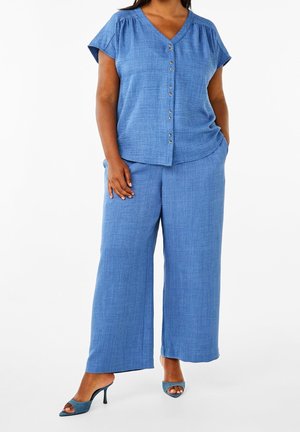 Kobieta w luźnym niebieskim topie z krótkim rękawem zapinanym na guziki i szerokich spodniach oraz niebieskich, otwartych, na obcasie butach typu mule na białym tle.