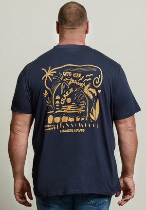 Mann trägt ein dunkelblaues T-Shirt mit gelber Grafik von Palmen, Sonne, Ozean und dem Text „Off the Beaten Track“ und „Chasing Waves“ auf dem Rücken.