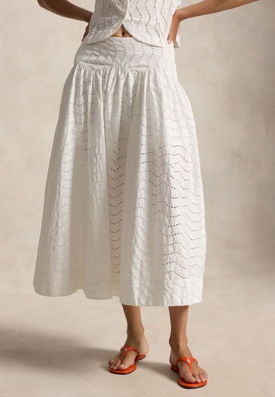 Femme portant une jupe midi blanche en œillet avec une taille froncée et des sandales orange à lacets, posant les mains sur les hanches devant un fond uni.