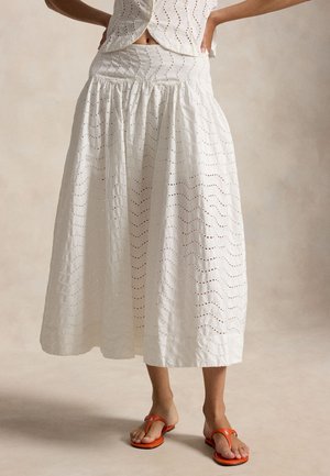 Femeie purtând o fustă midi albă cu broderie perforată și talie adunată, cu sandale portocalii tip șlapi, pozând cu mâinile pe șolduri pe un fundal simplu.