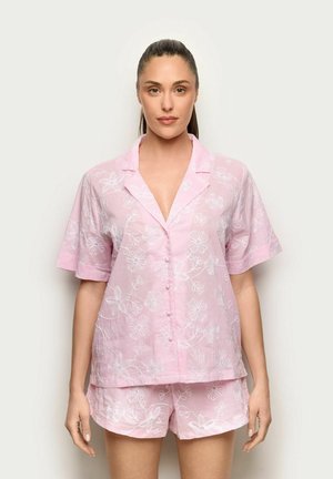 Femme portant une chemise à manches courtes rose clair avec boutons et un short assorti avec broderie florale blanche, debout devant un fond uni.