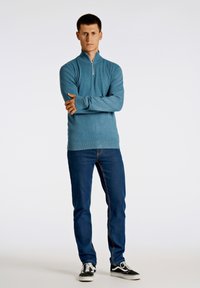 Ljusa blå dragkedjetröja med ståndkrage, ribbade mudd och fåll, kombinerad med mörkblå jeans och svarta sneakers, på en neutral bakgrund.