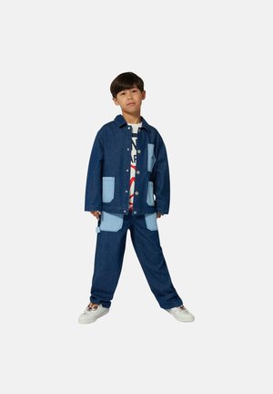 Veste en denim avec des poches bleu clair, associée à un pantalon assorti. T-shirt graphique blanc en dessous, porté avec des baskets blanches.