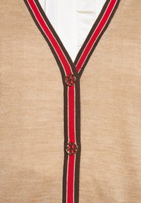 Beiger Cardigan mit V-Ausschnitt, der rot- und braun gestreifte Details entlang der Kanten und goldene Knöpfe mit einem eingravierten Design aufweist.