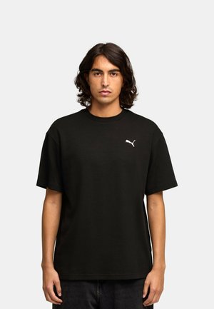 Młody mężczyzna z ciemnymi włosami do ramion, ubrany w czarną koszulkę Puma na jednolitym jasnoszarym tle.