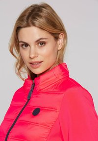 Padded red vest with high collar, featuring a zip closure and a solid black logo patch on the chest; fabric has a smooth, shiny texture.
