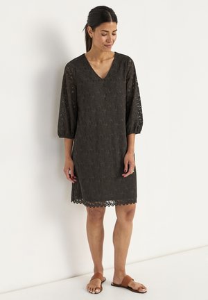 Robe en dentelle noire avec un décolleté en V, des manches bouffantes 3/4, un ourlet festonné et une texture transparente, associée à des sandales plates marron.