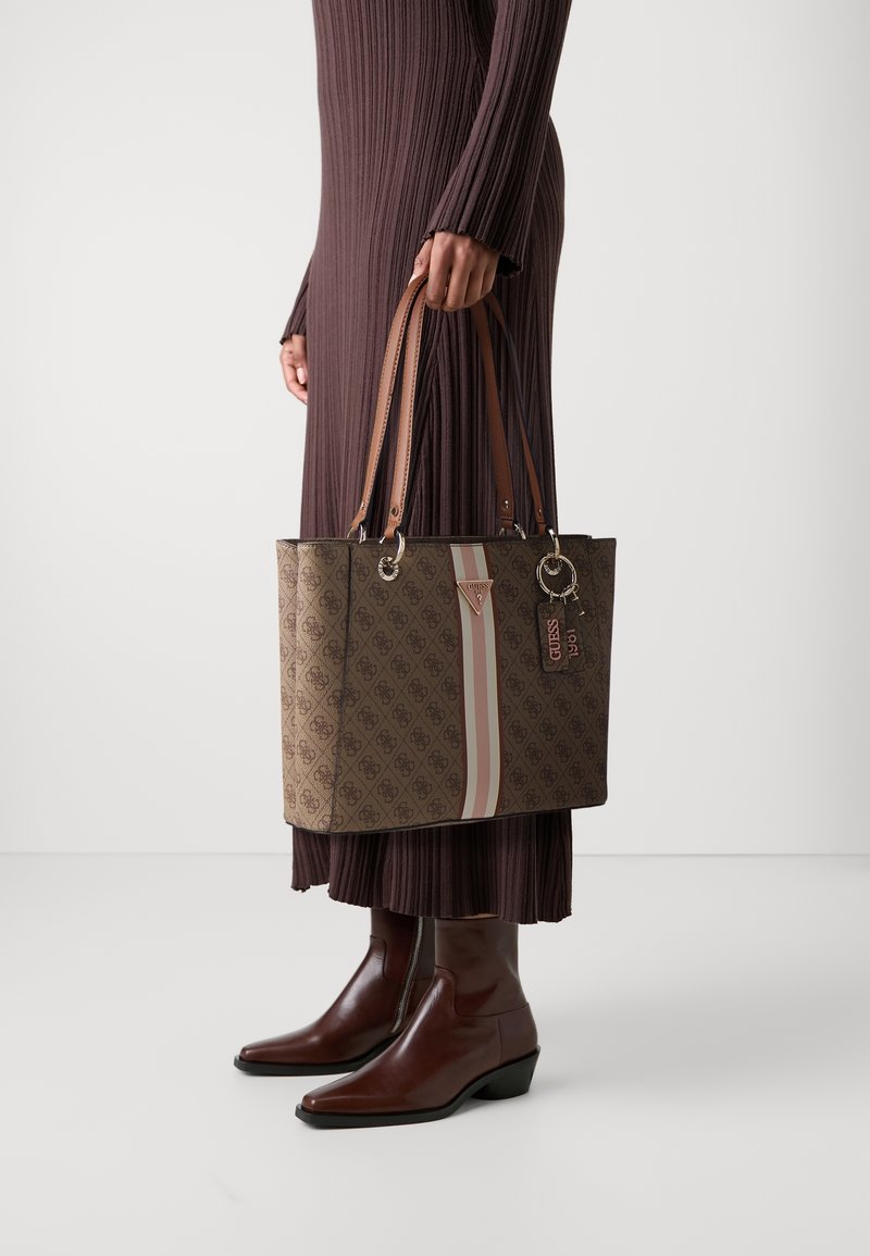 Personne en longue robe côtelée marron et bottines marron tenant un sac cabas marron à motifs avec des anses en cuir et des rayures verticales roses et blanches.