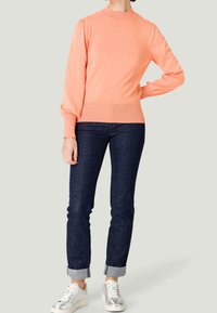 Koraalkleurige trui met lange mouwen en een scalloped halslijn, geribbelde manchetten en zoom, gecombineerd met donkere spijkerbroek en metallic sneakers.