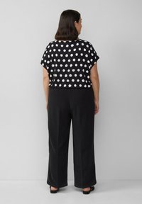 Vrouw met schouderlang zwart haar, gekleed in een zwart topje met korte mouwen met stippen en losse zwarte broek, staande tegen een effen muur.