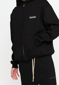 Sudadera con capucha negra con cremallera, hecha de tela suave con un bolsillo frontal, con logo "MULTIPLY APPAREL" en blanco. Incluye puños y dobladillo acanalados.
