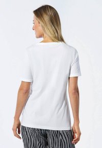 Weißes kurzärmliges T-Shirt mit rundem Halsausschnitt. Glatte Textur, lockere Passform, sichtbare Nähte und umgeschlagene Ärmel. Gemusterte schwarze Hose.