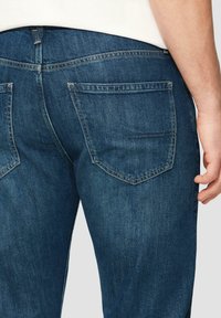 Jean en denim bleu foncé avec un design classique à cinq poches, présentant une taille mi-haute et une légère décoloration sur le tissu.