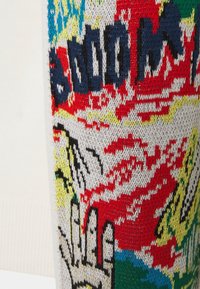 Gros plan sur un tissu tricoté coloré avec des motifs abstraits audacieux en rouge, jaune, bleu et noir, ainsi qu'un texte partiel sur un fond blanc cassé.