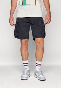 Black cargo shorts with side pockets, cotton blend material, relaxed fit. Paired with gray sneakers and white socks featuring red and blue stripes.