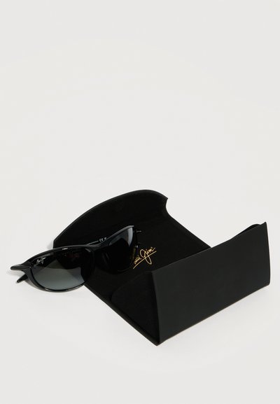 Μαύρα γυαλιά ηλίου cat-eye που ακουμπούν σε μια ανοιχτή μαύρη θήκη με χρυσή καλλιγραφική σήμανση, πάνω σε λευκή επιφάνεια.