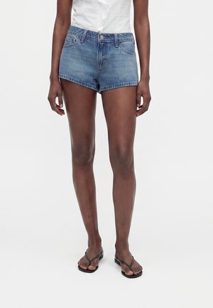 Personne portant un short en jean bleu taille haute, un haut blanc sans manches avec des volants et des sandales flip-flop noires, debout sur un fond blanc.