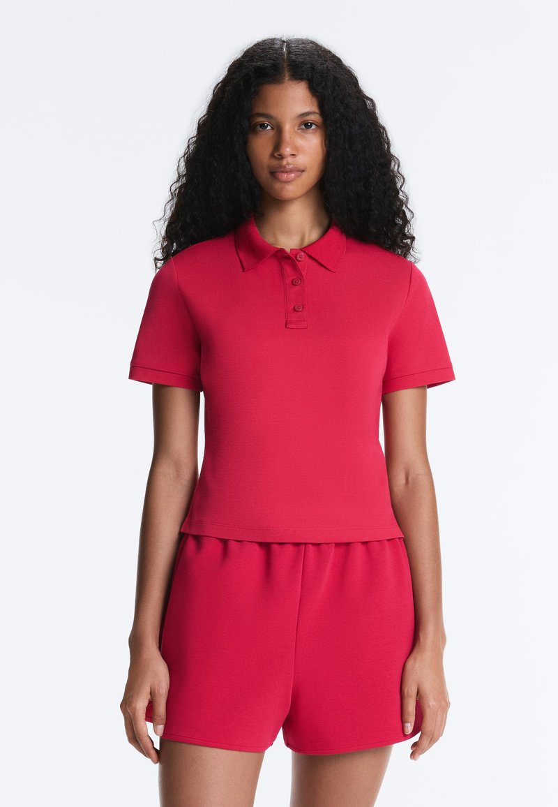 Femme aux longs cheveux bouclés portant un polo rouge à manches courtes et un short rouge assorti, debout devant un fond blanc uni.