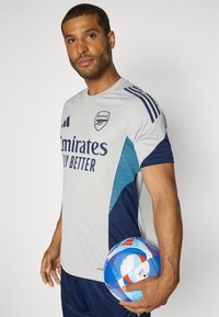 Grijze sportshirt met blauwe accenten, voorzien van het Arsenal-logo en de tekst "Emirates Fly Better". Houdt een multikleuren voetbal vast.