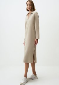 Robe pull beige avec un col, des manches longues et des fentes latérales. Réalisée en tissu texturé doux, portée avec des baskets blanches.