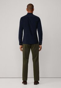 Navyblauw overhemd met lange mouwen, een kraag en knoopmanchetten, gecombineerd met olijfgroene broeken. De stof lijkt glad en gestructureerd.