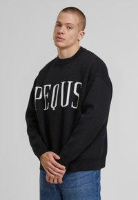 Svart genser med ribbestrikket hals, med fet hvit tekst "PEQUS." Laget av myk stoff, sammen med lyseblådenim jeans.