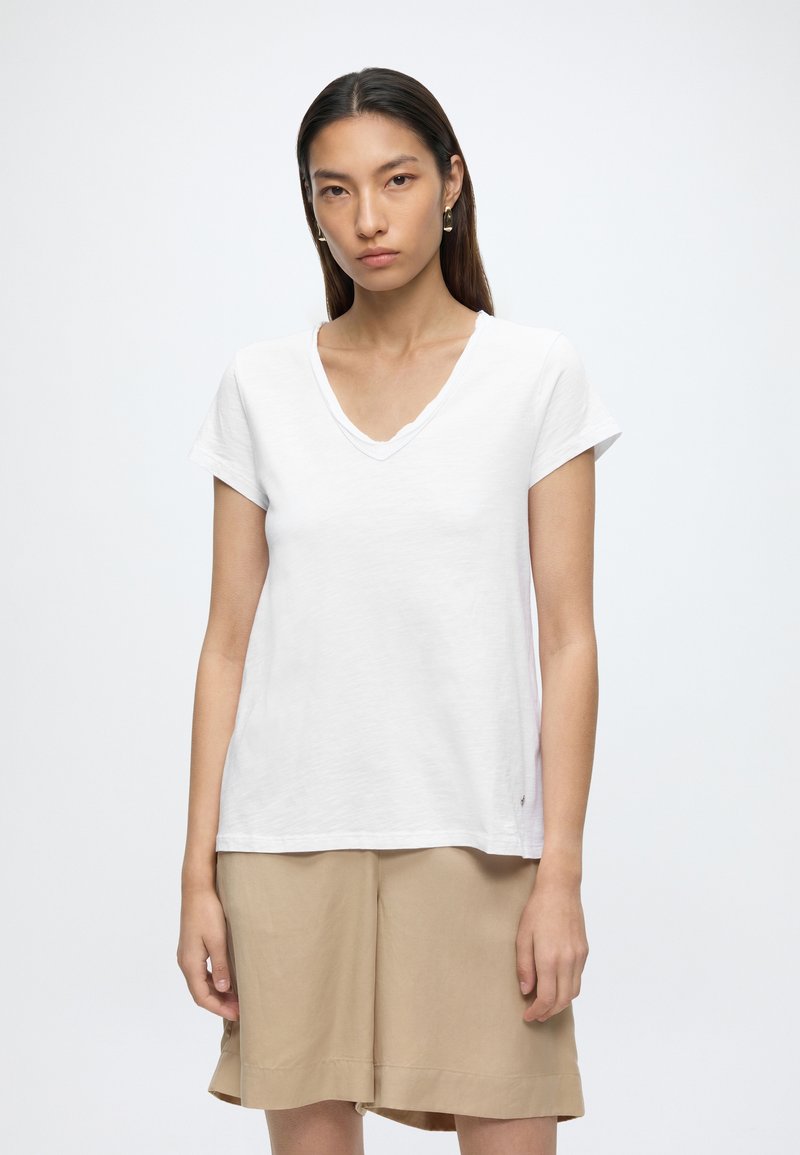 Jeune femme aux longs cheveux foncés porte un t-shirt blanc à col en V et un short beige, debout devant un fond clair uni.