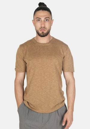 Uomo con capelli scuri raccolti in uno chignon, indossa una camicia marrone a maniche corte e pantaloni grigi, in piedi con una mano in tasca su sfondo bianco.
