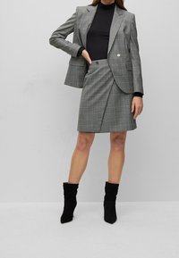 Grijs geruite blazer en rok set met zwarte coltrui. De blazer heeft een enkele knop; de rok heeft een wikkelontwerp. Inclusief zwarte slouch-boots.