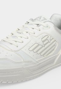 Baskets blanches avec lacets, détails perforés et un logo stylisé sur le côté, dotées d'une semelle texturée et d'un col rembourré.