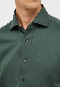 Groene gestructureerde shirt met een gestructureerde kraag, voorzien van zwarte knopen en een subtiel patroon, gezien vanaf de borst omhoog.