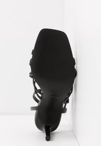 Sort højhælet sandal med tynde stropper, firkantet tå og struktureret sål, anlagt mod en hvid hjørne baggrund.