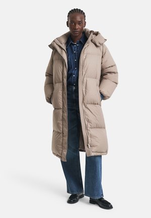 Beige Steppjacke mit Kapuze, ausgestattet mit einem gepolsterten Design und Druckknopfverschlüssen; getragen über einem dunklen Jeanshemd und einer weit geschnittenen Jeans.