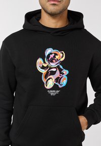 Svart hoodie med en färgglad björngrafik på framsidan, i ett texturerat tyg och med framficka. Logotypen är centrerad under motivet.