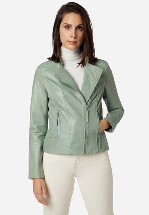 Giacca da biker in pelle verde menta con colletto a revers, zip asimmetrica e due tasche laterali. Indossata sopra un dolcevita bianco.