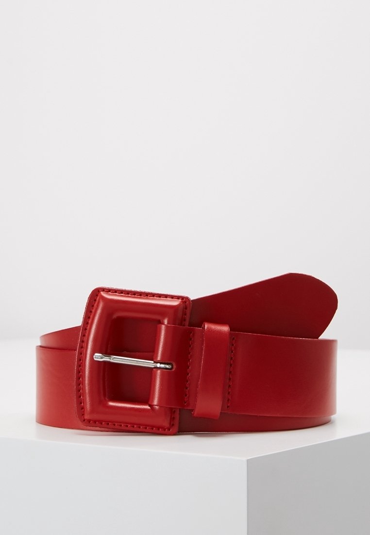 Ceinture en cuir rouge avec une large boucle rectangulaire et une pointe métallique, enroulée et posée sur un socle blanc sur un fond uni.