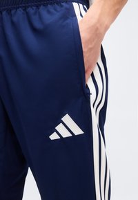 Χέρι που ακουμπά στην τσέπη σκούρου μπλε αθλητικού παντελονιού Adidas με λευκές τρεις ρίγες στο πλάι και λογότυπο στο μηρό.