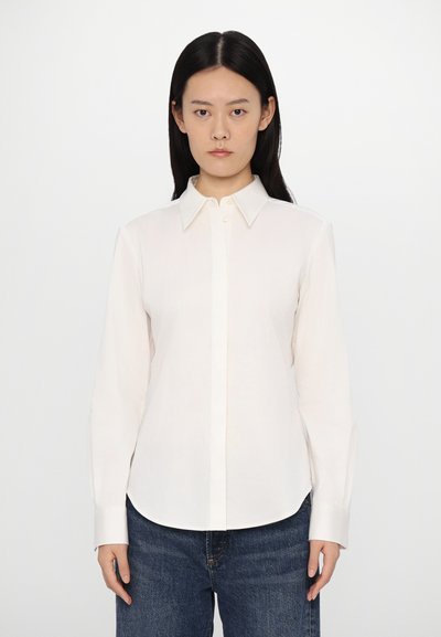 Chemise blanche à manches longues, en tissu léger, avec un col pointu et des poignets classiques, associée avec un jean en denim bleu.