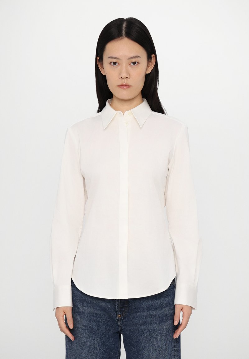 Long-sleeve, white button-up shirt in lightweight fabric with a pointed collar and classic cuffs, paired with blue denim jeans.