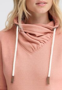 Felpa rosa chiaro con un collo piegato e arricciato. Presenta un scollo triangolare e cordini bianchi in contrasto con accenti marroni.