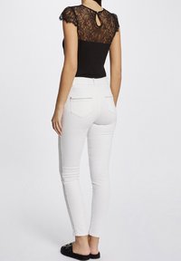 Top de encaje negro con mangas cortas y apertura en la espalda, combinado con jeans blancos ajustados que tienen bolsillos traseros y cremalleras. Zapatos de deslizamiento negros.
